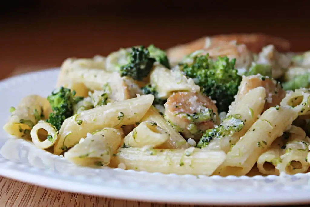 A close-up image of creamy chicken broccoli ziti on a white plate, with steamed broccoli, tender chicken pieces, and a sprinkle of Parmesan cheese on top, presented on a wooden table.